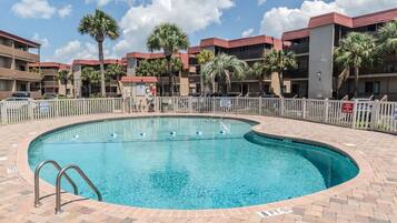 A swimming pool with a brick border, surrounded by a white fence, and adjacent to a multi-story building with balconies.