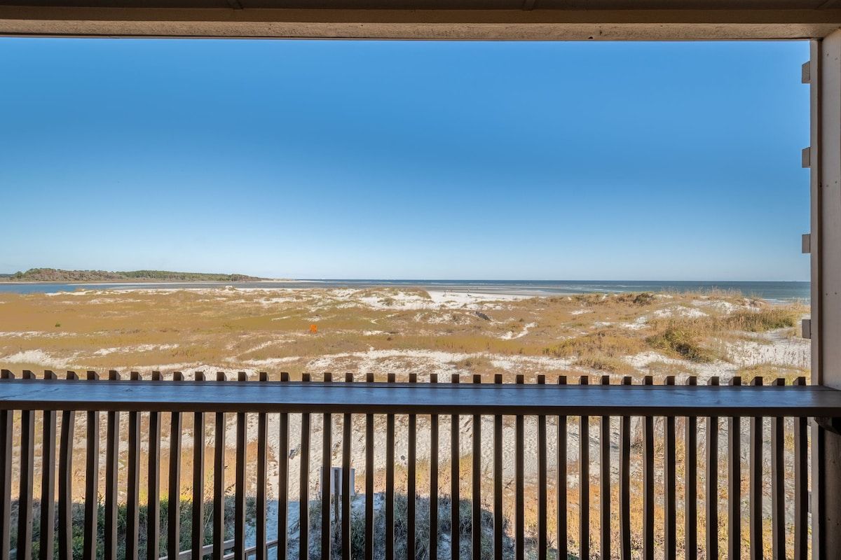 A view from a balcony with a railing, overlooking a beach with sand dunes and a clear sky.