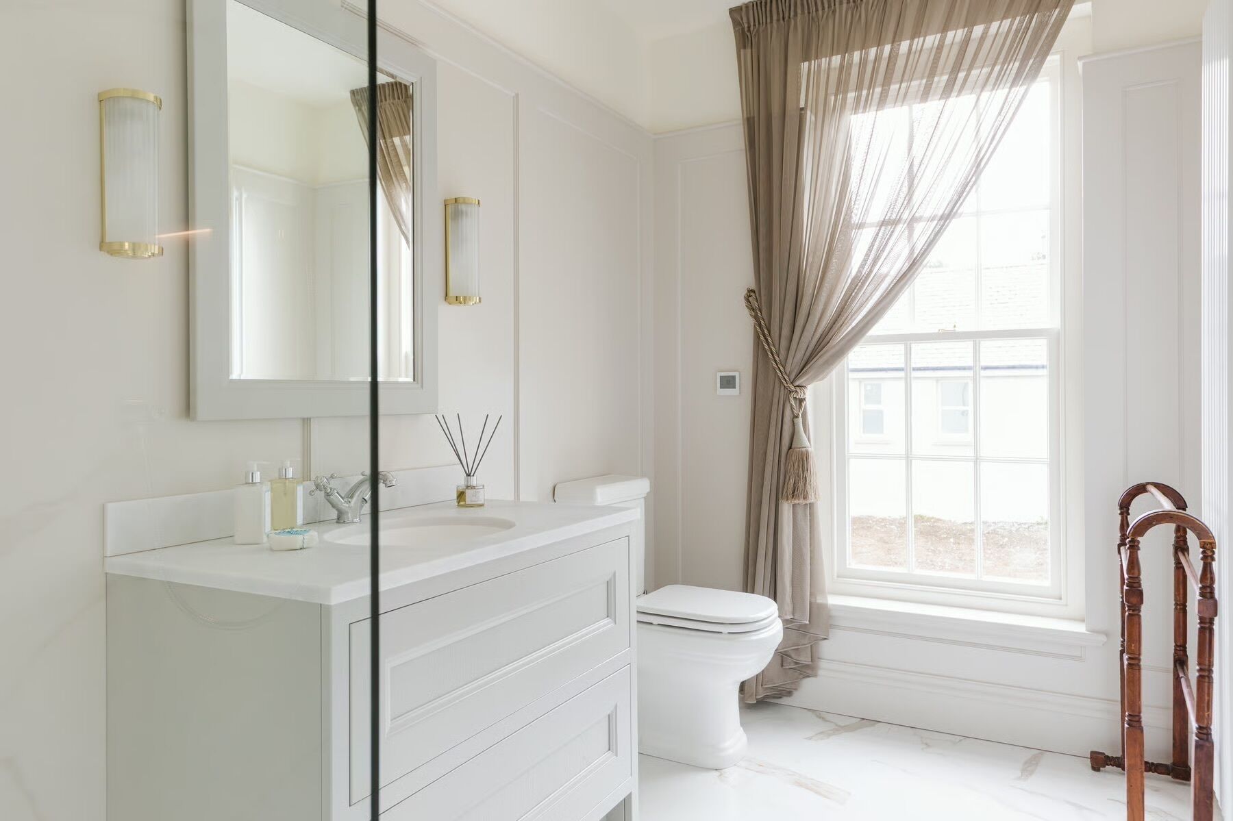 Un baño con tocador blanco, inodoro, espejo grande y ventana con cortinas.
