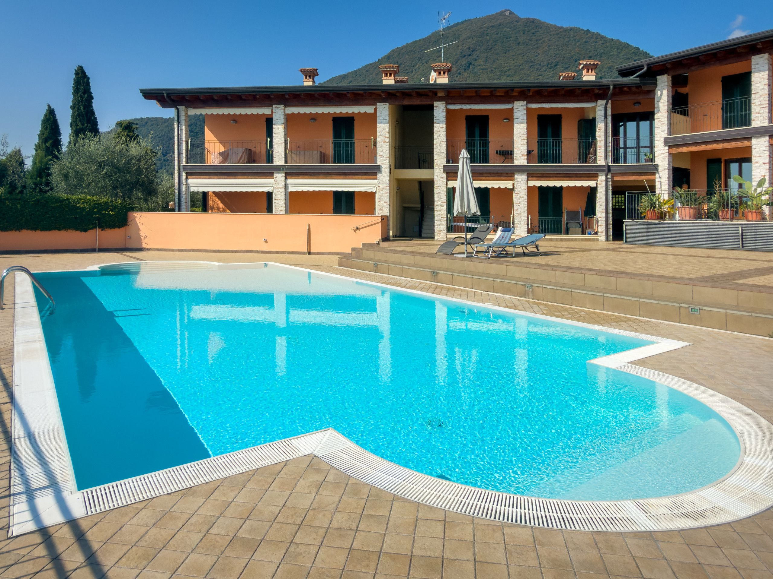 A swimming pool with a tiled deck, a building with balconies, and a mountain in the background.