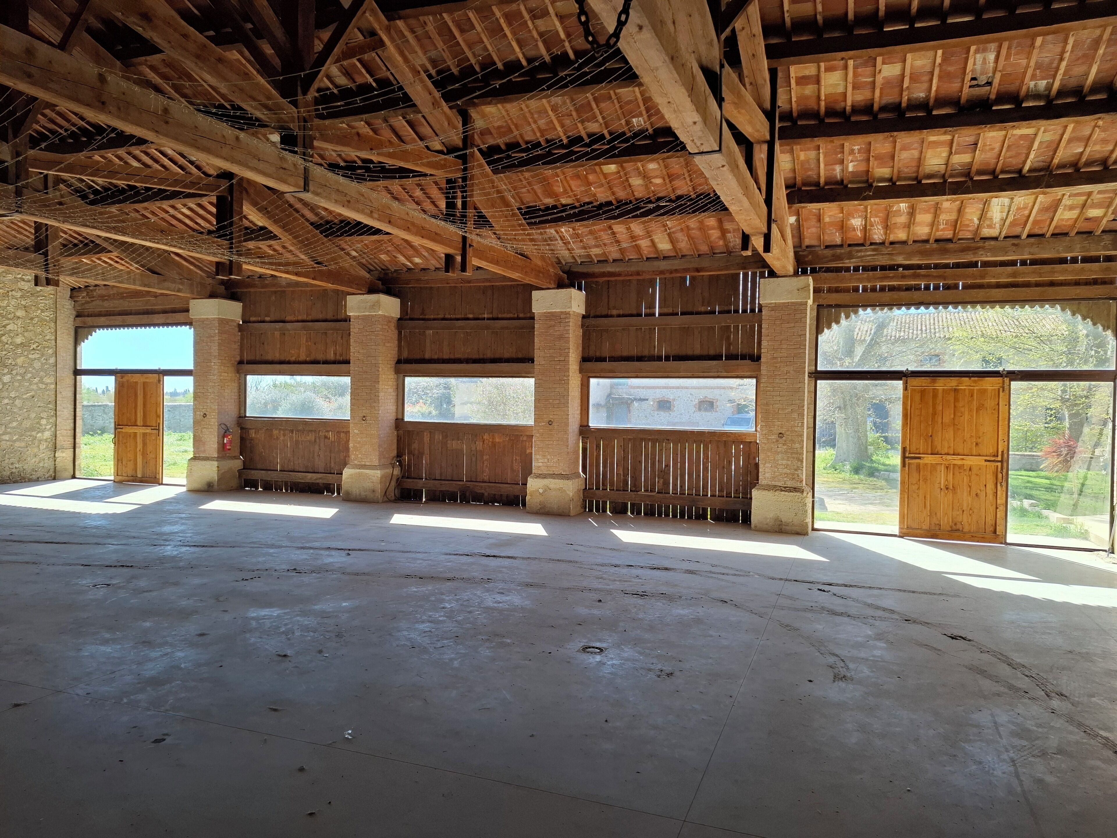 Ein Innenraum mit hoher Decke, Holzbalken, großen Fenstern und einem Betonboden.