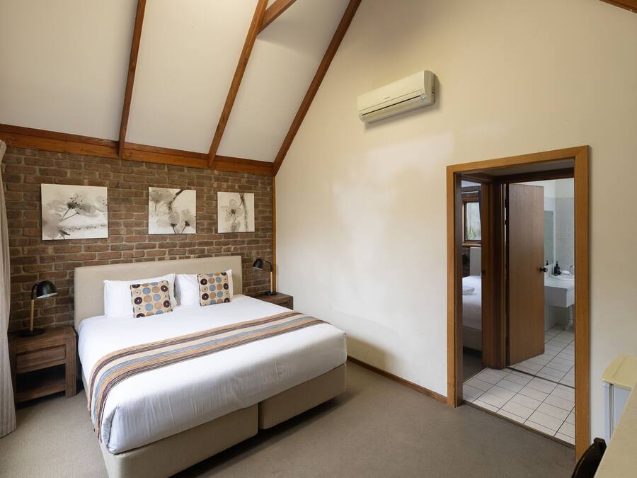 Un dormitorio con pared de ladrillo, cama con cubrecama a rayas, mesita de noche con lámpara y puerta que da al baño.