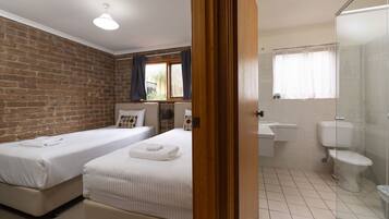 A hotel room with two beds, a brick wall, a window with curtains, a bathroom with a toilet and sink, and a shower area.