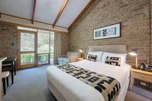 A hotel room with a large bed, a desk, a chair, and a balcony with a view of trees.