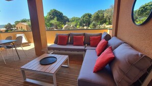 Een zonnig terras buiten met een houten tafel, een comfortabele bank met rode kussens en uitzicht op een natuurlandschap.