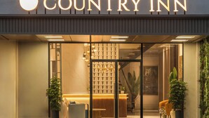 The exterior of a Country Inn hotel with a modern design, featuring a prominent illuminated sign, a reception area visible through the glass doors, and potted plants around the entrance.