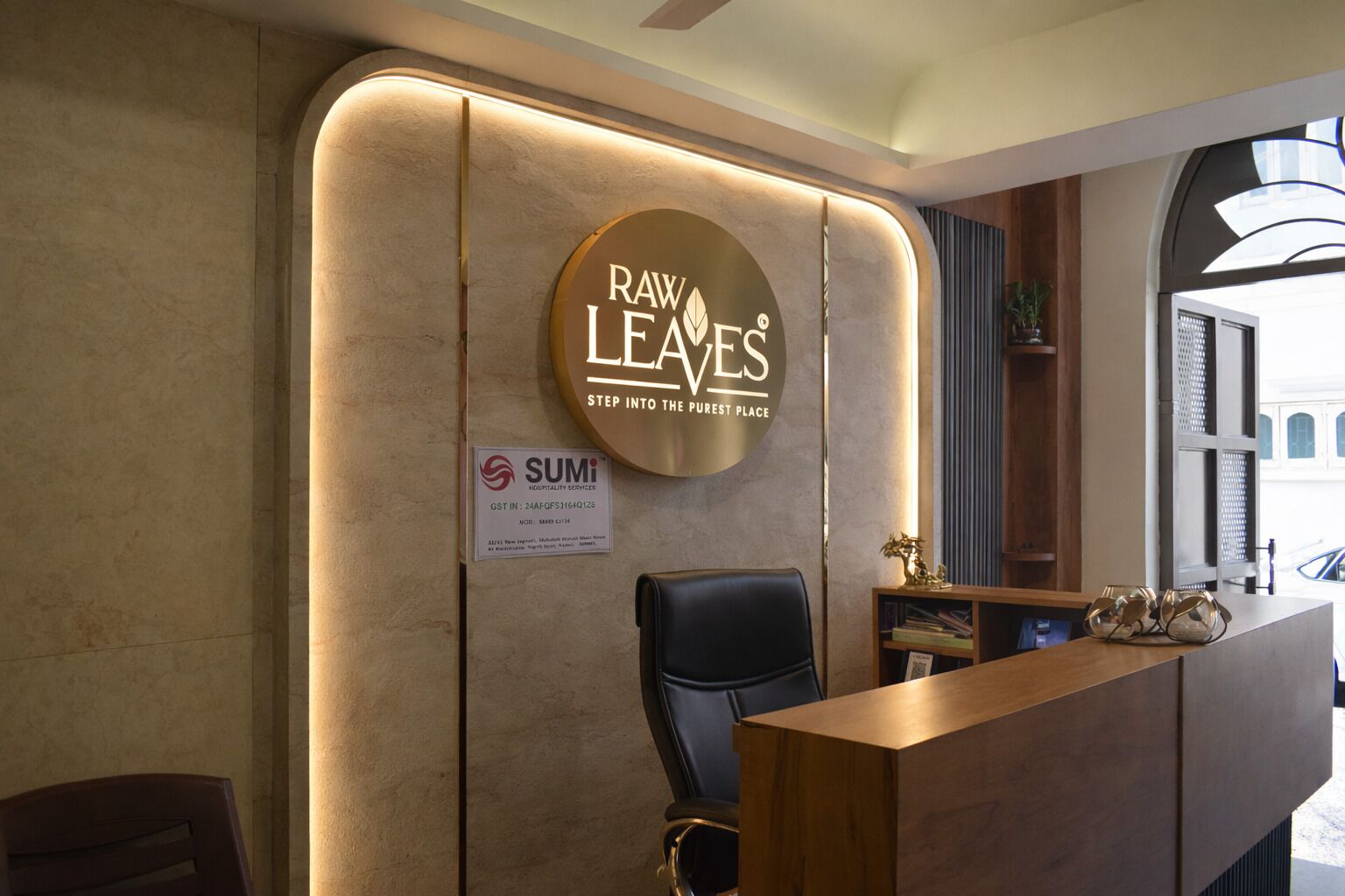 Ein Empfangsbereich mit einem Holztisch, einem schwarzen Bürostuhl und einem runden Schild mit dem Branding 'Raw Leaves'. Auch ein Schild mit 'SUMI' ist sichtbar.