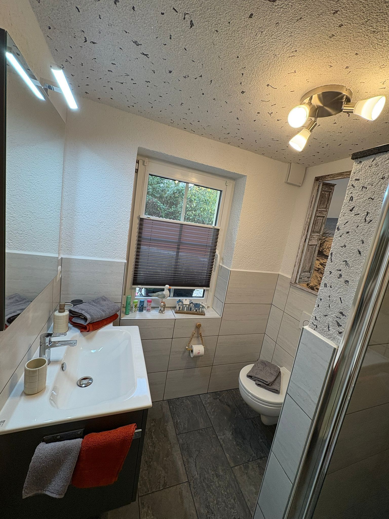 A bathroom with a white sink, mirror, and window with blinds. There is a toilet and a shower area.