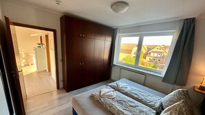 Ein Schlafzimmer mit Bett, Kleiderschrank, Fenster mit Blick auf Häuser und einer Tür zu einem Badezimmer.