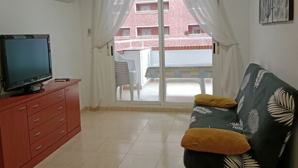 Ein Wohnzimmer mit Fernseher auf einem roten Schrank, einer gelb gekissenen Couch und einer Glastür zum Balkon.
