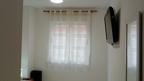 Ein Zimmer mit Deckenventilator, einem Fenster mit weißen Vorhängen und einem an der Wand befestigten Fernseher.