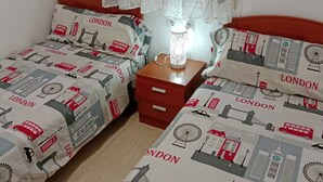 Ein Zimmer mit zwei Betten, jedes mit einem London-Motiv auf der Bettdecke, einem Nachttisch mit Lampe und einer roten Kommode.