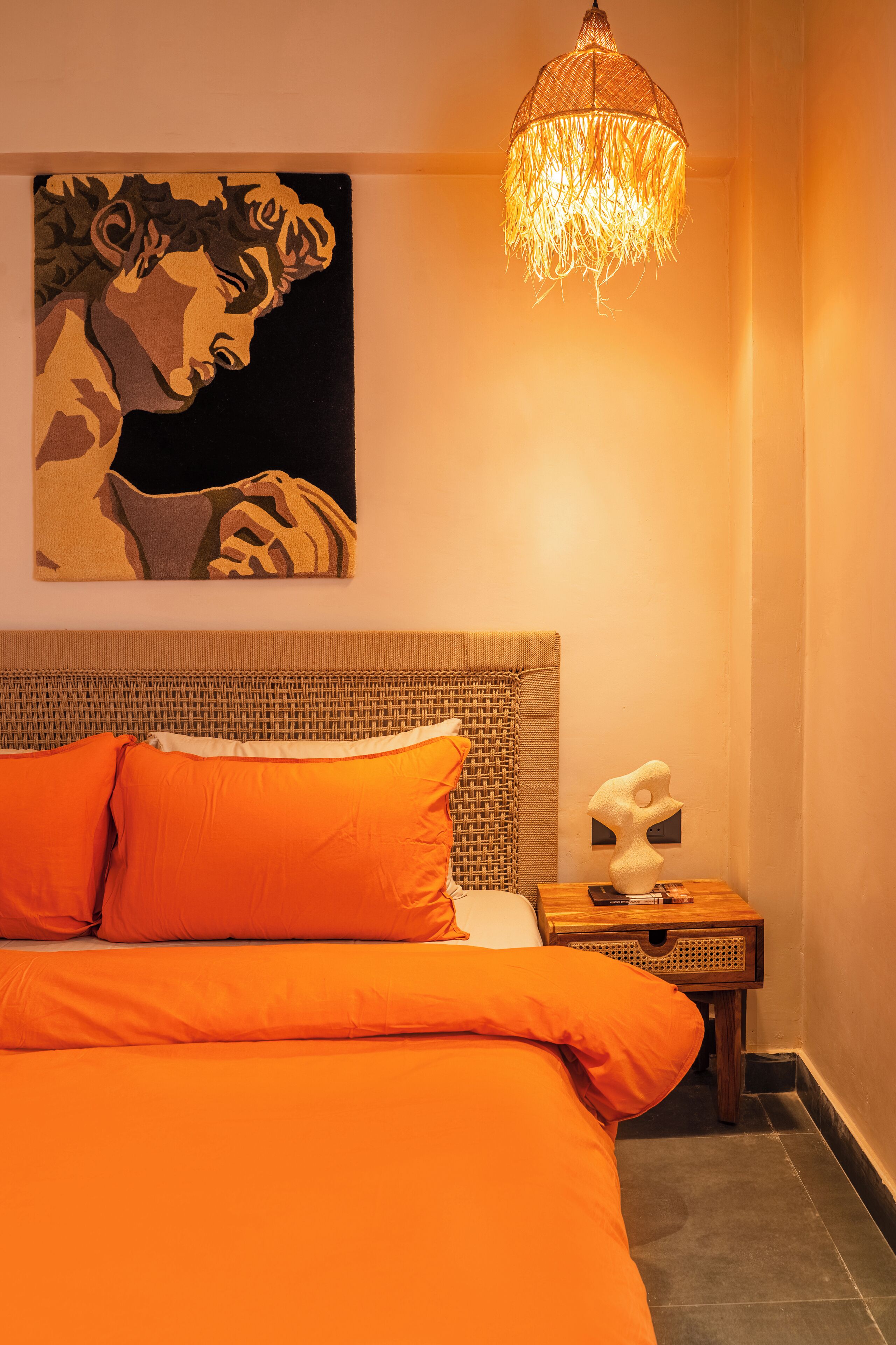 A bedroom with an orange bedspread, a woven headboard, a wall-mounted painting, a unique chandelier, and a small wooden nightstand with a sculpture.