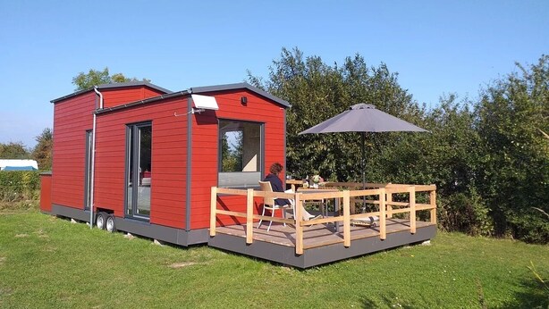 Une maison mobile rouge, dotée d’une terrasse et d’une table, entourée de verdure.