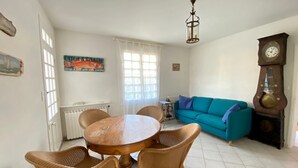 En matplats med ett runt träbord och flätade stolar, en turkosfärgad soffa, en stor väggklocka och ett fönster med gardiner.