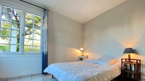 Ett sovrum med en säng, två sänglampor, ett fönster med gardiner och ett klinkergolv.
