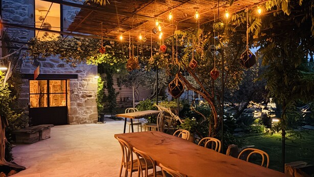 An outdoor dining area with a stone wall, hanging lights, and greenery. - LA MAISON BRUNO DI SALVIO (Les Vans)