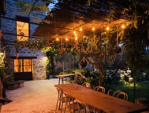 An outdoor dining area with a stone wall, hanging lights, and greenery. - LA MAISON BRUNO DI SALVIO (Les Vans)