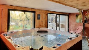 A hot tub in a wooden room with large windows, a wooden ceiling, and a chair by the window. - LA MAISON BRUNO DI SALVIO (Les Vans)