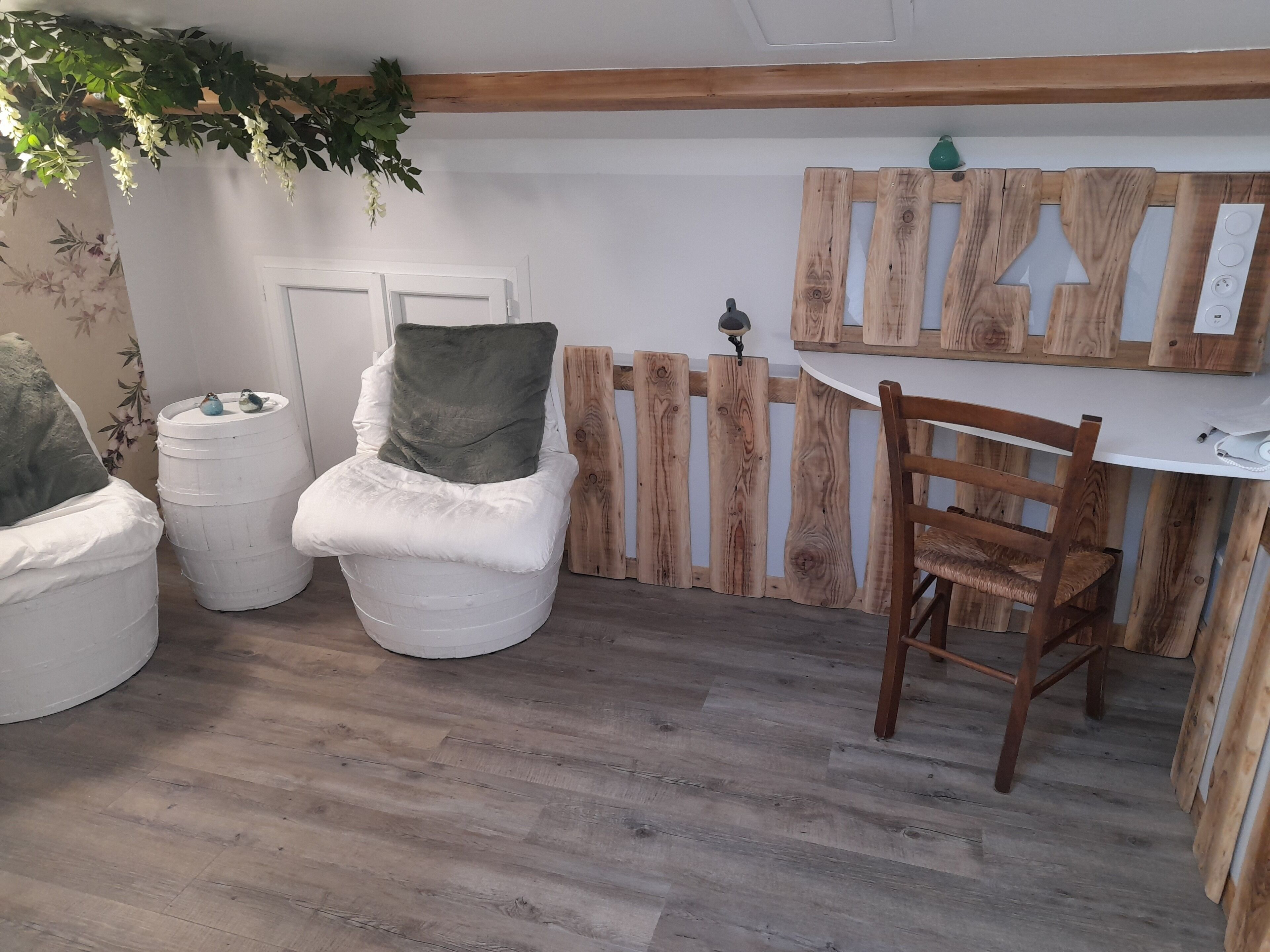 A cozy room with a wooden floor, a white cushioned chair with a green pillow, a wooden chair, and a white table with a wooden backdrop.