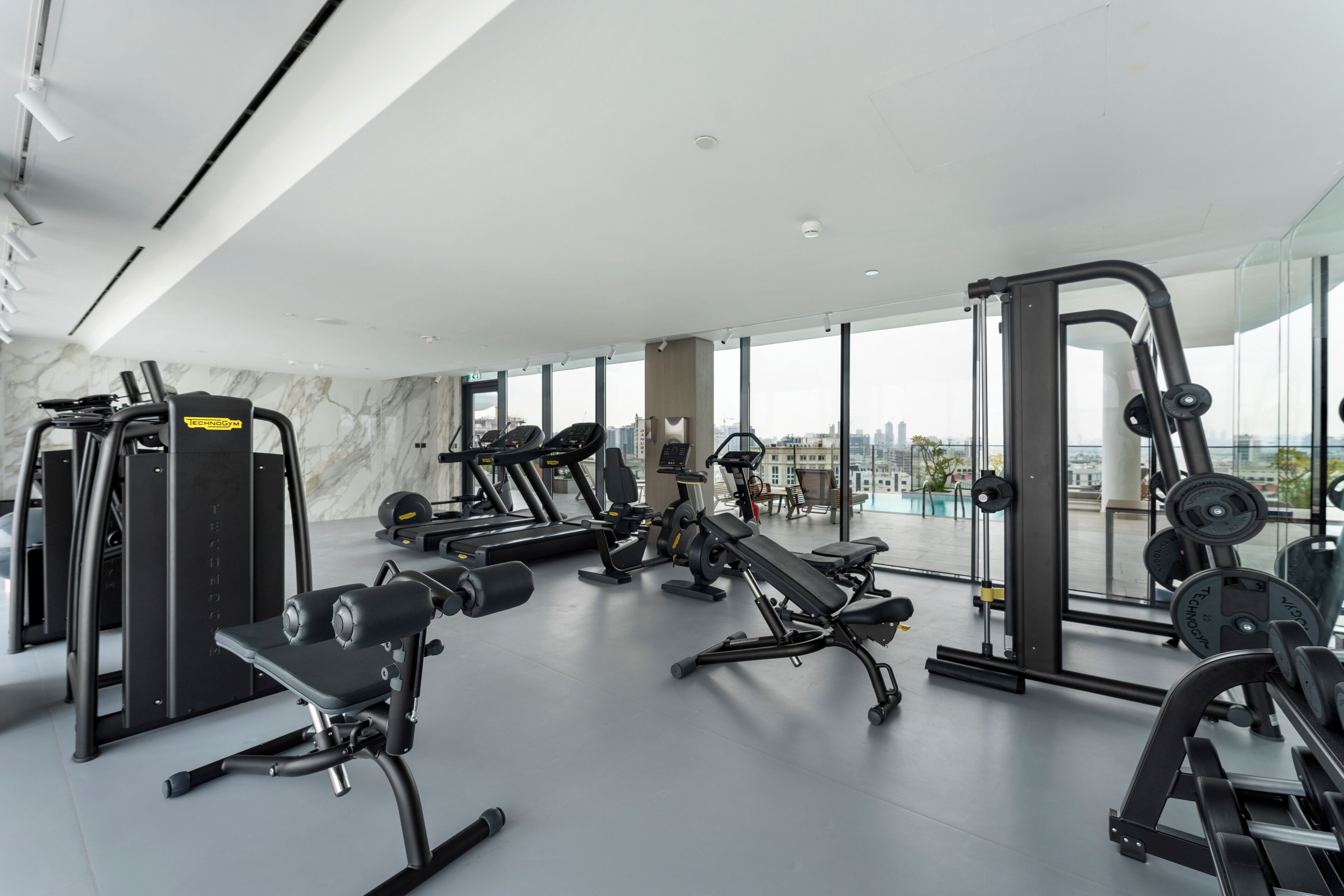 Un gimnasio moderno con diversos equipos de ejercicio, ventanas amplias y vistas a la ciudad.