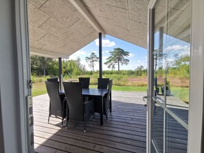En takförsedd uteplats med ett bord och stolar, ett trädäck och utsikt över en gräsyta med träd.