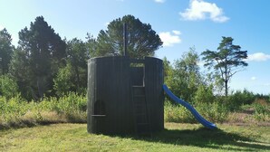 Une structure de jeu cylindrique avec un toboggan, entourée d’herbe et d’arbres.