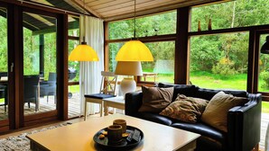 A cozy living room with a black leather sofa, a wooden coffee table, and a dining area with a white table and chairs. Large windows offer a view of a lush garden. - Charming Holiday Home With Spa and Sauna (Læsø)