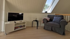 A modern living room with a sloped ceiling, a flat-screen TV on a white stand, a grey sofa with patterned cushions, and a small black side table. - Modern Holiday Home With Nature Views in Læsø (Læsø)