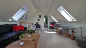 Een ruime loft met een hoge plafond, dakramen, een zwarte leren bank, een houten salontafel en een grijze fauteuil.