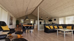 A spacious living area with a high ceiling, a dining table, black leather sofas with yellow cushions, and a wooden floor. - Panoramic Views and Luxurious Comfort on Læsø Island and Sleeps up to 16 Persons (Læsø)