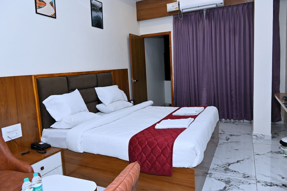 Une chambre d’hôtel avec un grand lit, une tête de lit en bois, du linge de lit blanc, un plaid rouge, une table de chevet en bois avec un téléphone et des bouteilles d’eau, et une salle de bain visible à travers une porte ouverte.