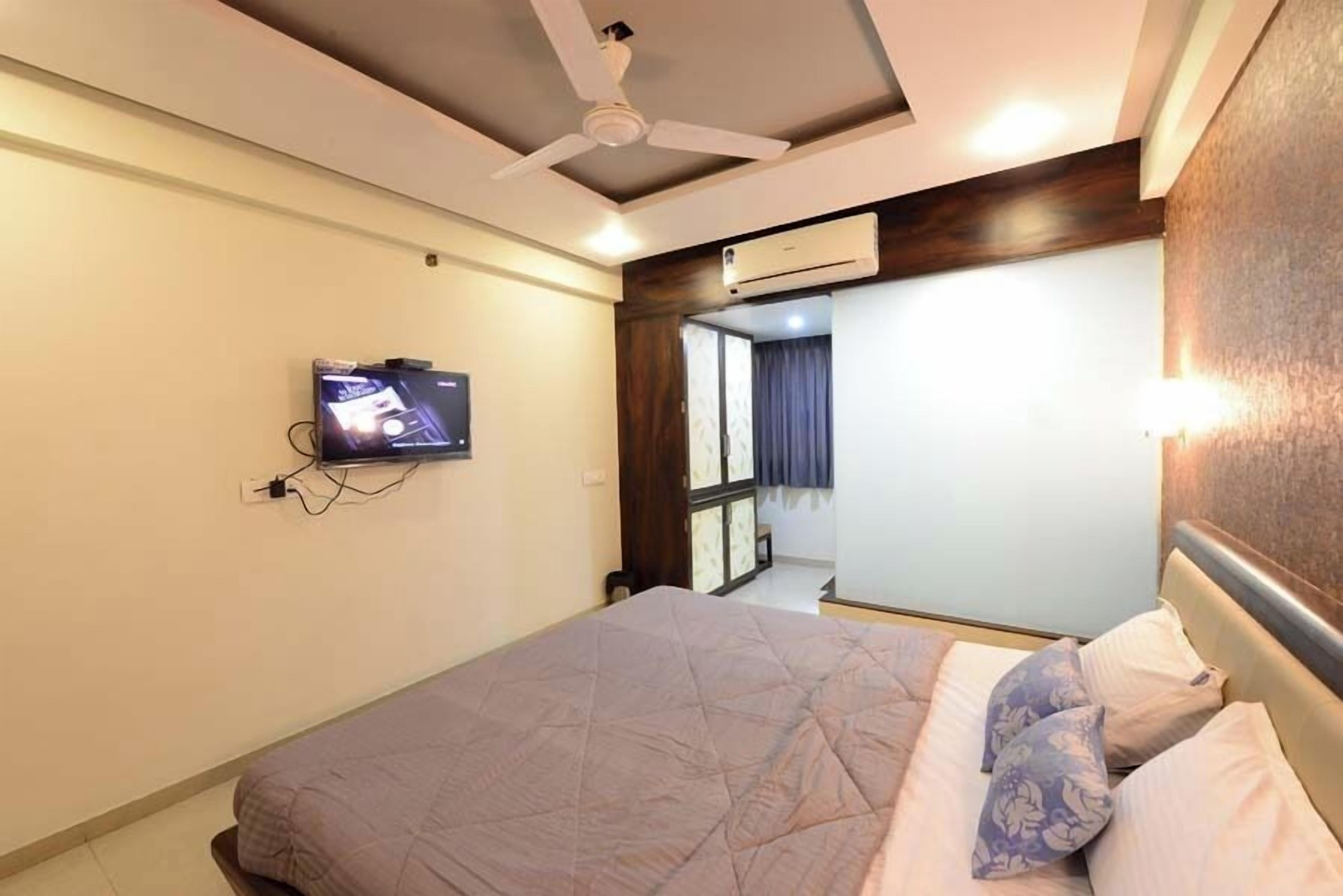 A hotel room with a bed, a ceiling fan, an air conditioning unit, and a television mounted on the wall.