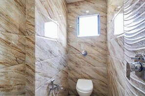 A bathroom with marble walls, a toilet, a shower, and a window. - Hotel Manila (Ahmedabad)