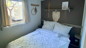 A bedroom with a bed, a window with curtains, a wall-mounted shelf with a picture, and a headboard with a decorative item. - Comfortable mobile home in the south of France - perfect family vacation near the beach (Vias)