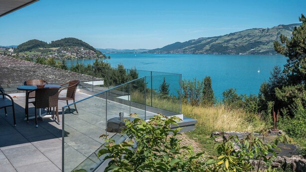 A terrace with a glass railing overlooking a lake, wicker furniture, and a view of hills. - Lake & Mountain View Villa | 15min to Interlaken (Spiez)