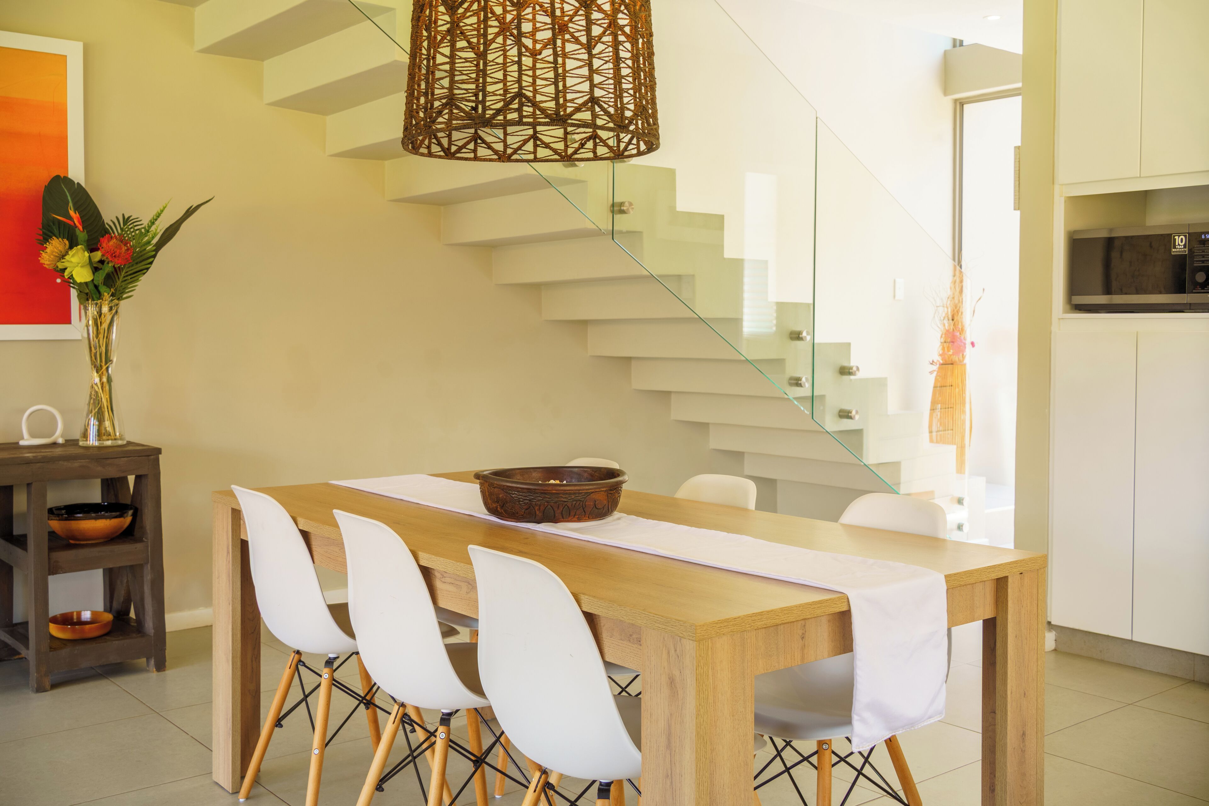 Un espace repas moderne avec une table en bois, des chaises blanches, un bol décoratif et un luminaire tressé.