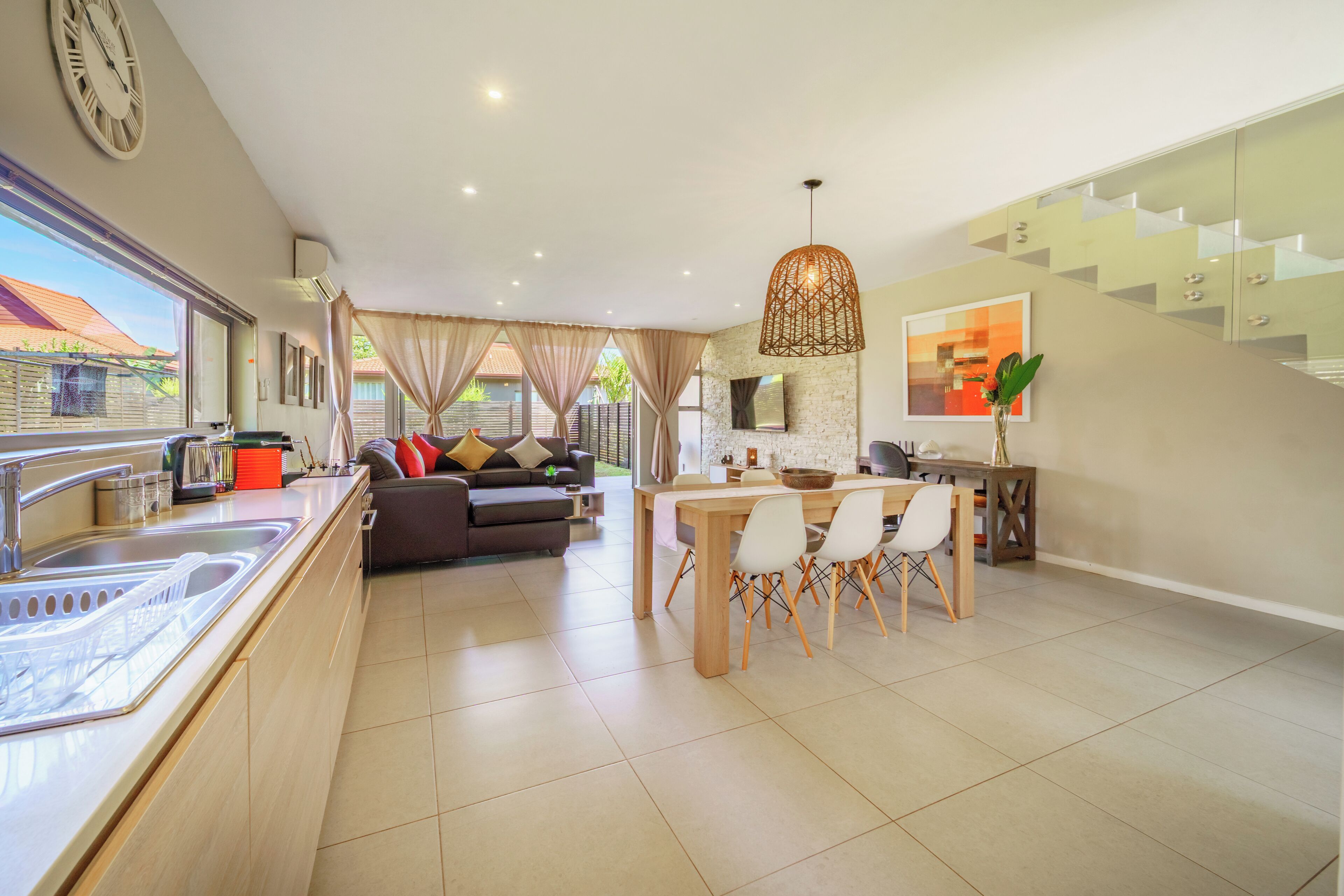 A modern kitchen with a dining area, a staircase, and a living room with a sofa and curtains.