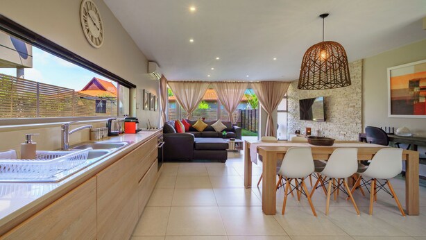 A modern kitchen with a dining area, a living room with a sofa and curtains, and a large window with a view of the outside. - Modern 3-Bed Home with Pool Access, Ballito (Ballito)
