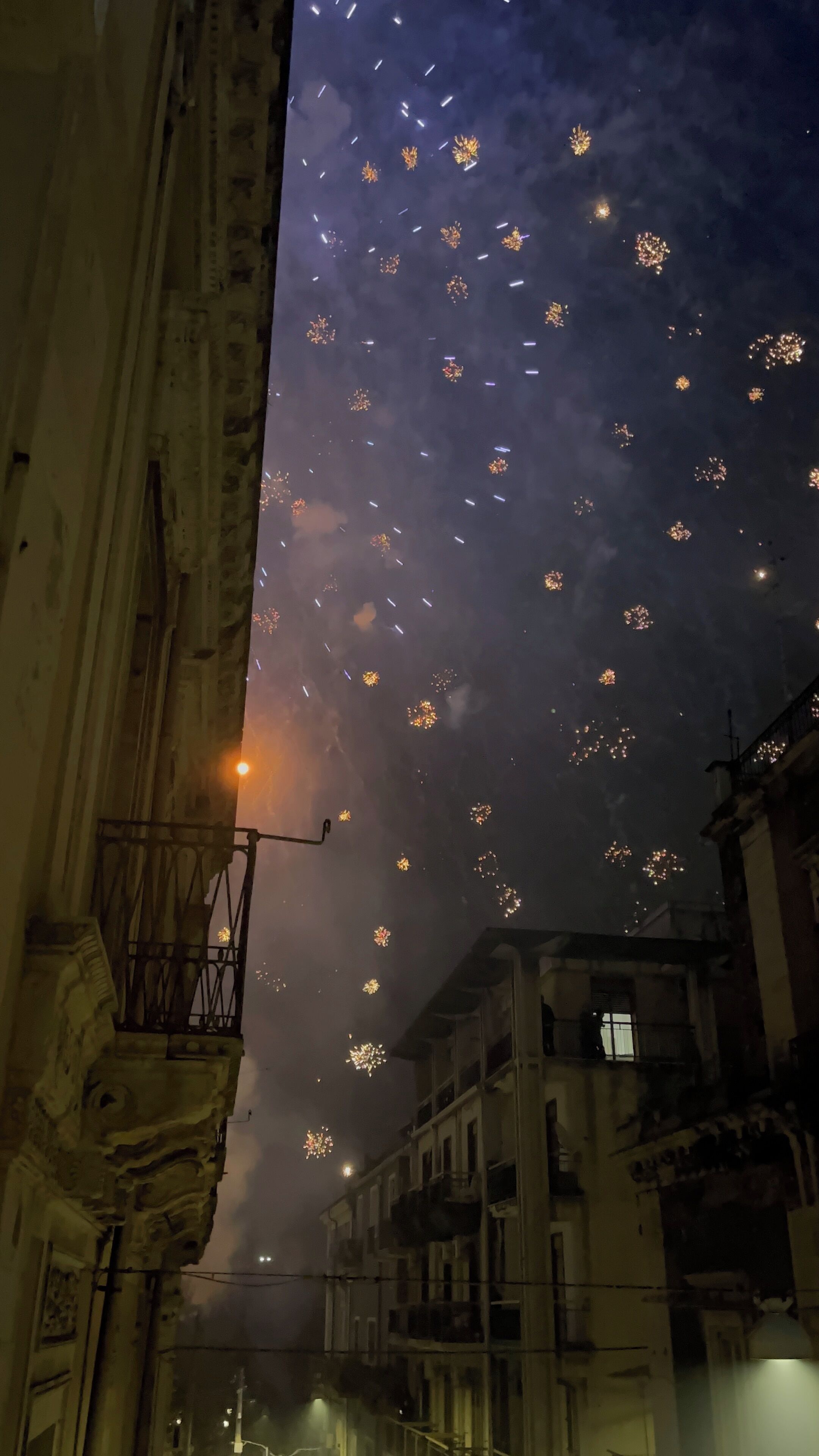 Une vue nocturne d’une rue bordée d’immeubles, avec des feux d’artifice dans le ciel.