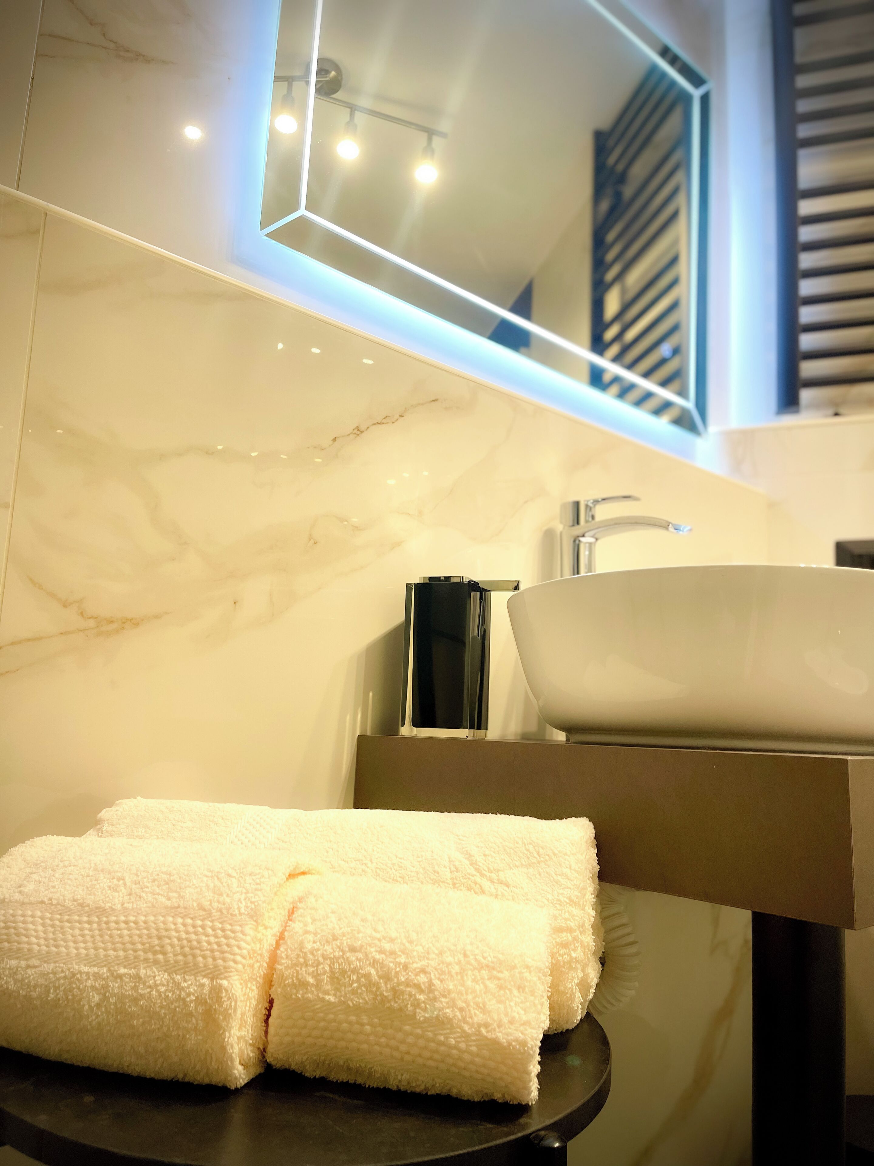 Un baño con pared de mármol, un lavabo blanco, una toalla sobre una bandeja negra y un espejo con luces.