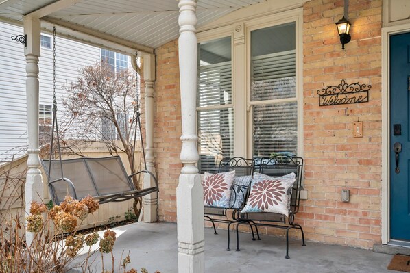 A porch with a swing chair, a bench with decorative pillows, a 'Welcome' sign, and a blue door.