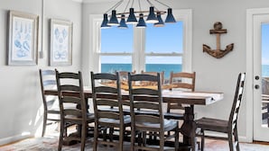 Un comedor con una mesa y sillas, una lámpara colgante, obras de arte enmarcadas y vista al océano.