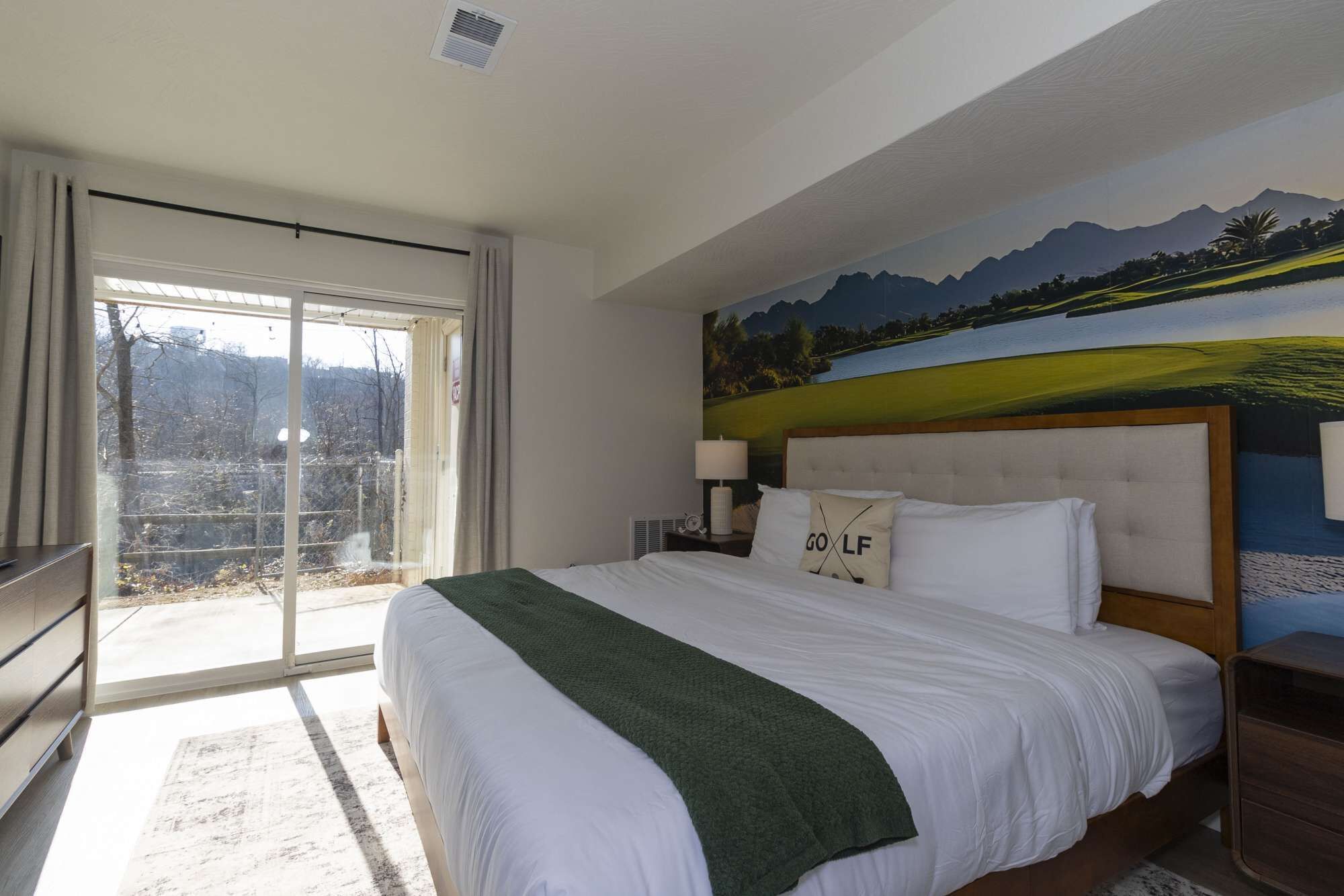 Une chambre d’hôtel avec un grand lit, une vue sur l’extérieur grâce à une porte coulissante, un tableau représentant un terrain de golf accroché au mur et une table de chevet avec une lampe.