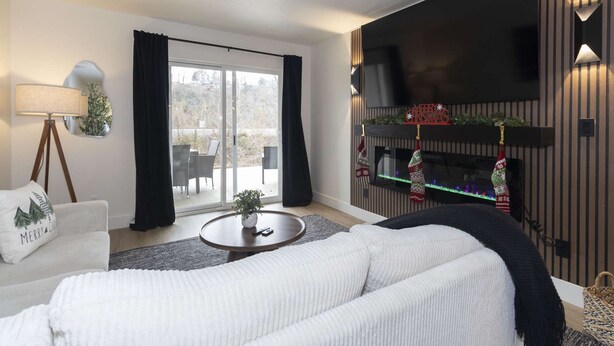 Una sala moderna con sofá, mesa de centro, lámpara de pie, chimenea con adornos navideños y un ventanal con cortinas.