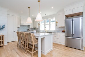A modern kitchen with white cabinetry, a stainless steel refrigerator, and a central island with bar stools. - Relaxin @ The Roses II - Brand New Home with Amazing Sound Views (Surf City)