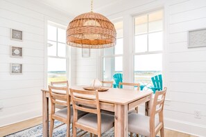 A dining area with a wooden table and chairs, a woven pendant light, and a view of the outdoors through large windows. - Relaxin @ The Roses II - Brand New Home with Amazing Sound Views (Surf City)
