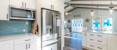 A modern kitchen with white cabinetry, stainless steel appliances, and a view to a beach house.