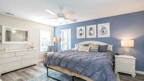 Ein Schlafzimmer mit Bett, Nachttischen, Kommode, Deckenventilator und Kunstwerken an der Wand.