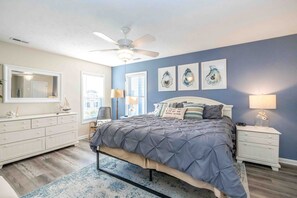Ein Schlafzimmer mit Bett, Nachttischen, Kommode, Deckenventilator und Kunstwerken an der Wand.